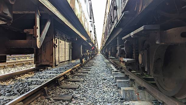 Between two trains beneath carriage viewing train car, wheels, concrete sleepers, rusting railroad tracks. Rusty derelict undercarriage of obsolete public transport network railway line. Rail transportation commuting journey travelling straight to horizon. Deserted dilapidated carriages, rails, sleeper on national rail transit. Travel or commute apocalyptic imagery of abandoned industrial railcar on train track.