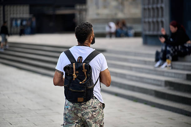 Young man with a backpack approaches a historic stone building in daylight, vertical frame, dynamic street composition for travel, education, commuting, or tourism campaigns.