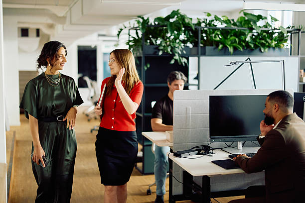 istock Two professional women chatting while walking together in an eco friendly office with colleagues in background 2234284572