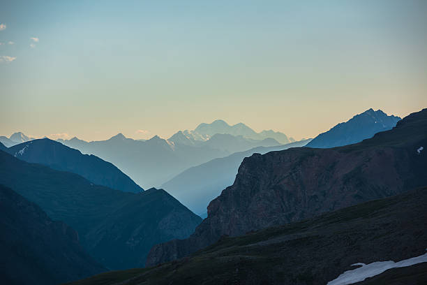 istock Deep gorge between layered silhouettes of sharp rocky ridge spurs under huge snow-capped mountain peak in golden blue gradient sky. Sheer crags of mount valley under giant ice top of gold sunset color 2234286952