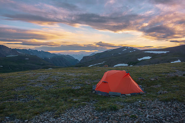 Orange tent on grassy stony pass with view to mountain range silhouette far away under clouds in vivid sunset color sky. Scenic alpine landscape with red tent in mountains under colorful cloudy sky.