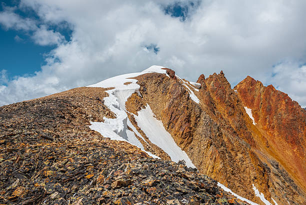 istock Snow-capped mountain ridge with cornice and red sharp rocks on top in sunlight. Snow cap and shiny pointy peak of gold color. Snow dome and colorful rocky peaked top. Sunny cloudy high mountains view. 2234288307