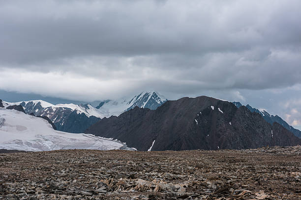 istock Dramatic misty top view from stone pass to big glacier tongue among sharp rocks and large snow-capped pinnacle in rainy low clouds. Dark atmospheric mountain silhouettes in rain in gray cloudy sky. 2234289473