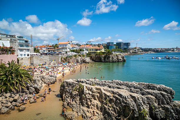 19 August 2025 Cascais, Portugal. Tourists relax on a Praia da Rainha in Cascais, Portugal, surrounded by rocky cliffs and turquoise waters. A lively summer atmosphere with swimmers, sunbathers, and boats under a clear blue sky on the Atlantic coast.