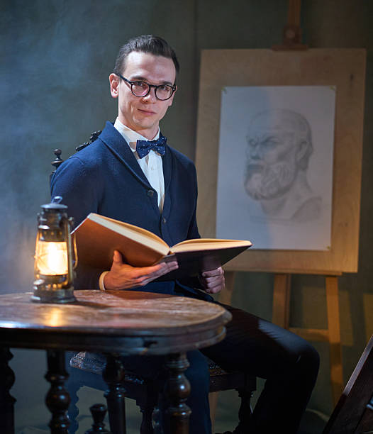 A attractive young man dressed in formal clothes, in bow tie and in glasses is sitting at old round table. He is holding open big book. Shooting in studio decorated in retro style