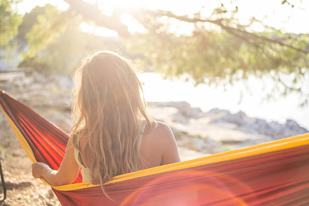 Young woman with long hair sitting in a red and yellow hammock near a lake during sunset. Relaxing outdoor leisure time by water. Peaceful nature and calm evening scene.