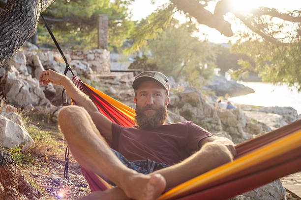 Bearded man wearing cap resting in colorful hammock tied to tree. Casual outdoor relaxation by rocky coast with sunlight filtering through trees. Leisure time in nature near water.