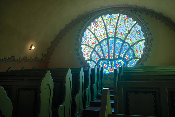 Large colorful circular stained-glass rose window framed by soft glow. Wooden pews at foreground. Benches in synagogue