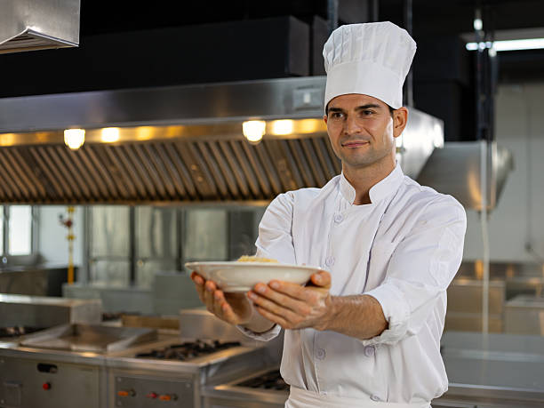 istock Smiling chef in uniform and hat proudly presents a finished dish while standing inside a modern stainless steel restaurant kitchen 2234312962