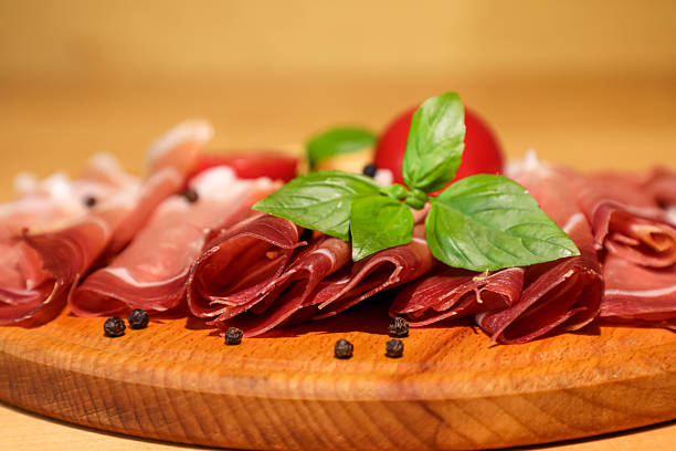 Delicate prosciutto slices served on a wooden board with ripe tomatoes, fragrant basil, and whole black peppercorns. Perfect for gourmet food and Italian cuisine themes.