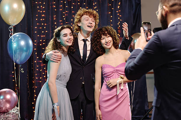 Group of three teenagers, two Caucasian girls and one Caucasian boy, standing together smiling while posing for smartphone photo at formal event with balloons and string lights in background