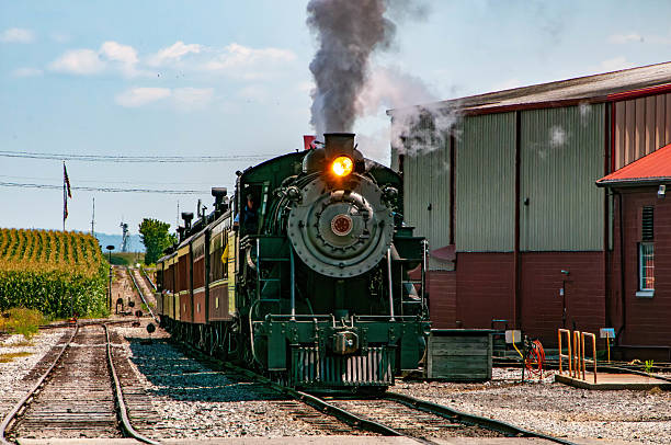 A steam locomotive releases plumes of smoke while departing from a quaint station. Sunlight illuminates the surrounding fields, highlighting a picturesque rural setting.