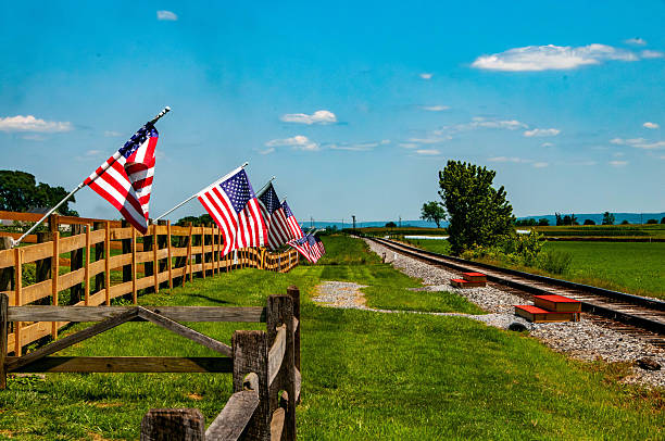 Brightly colored American flags flutter along a wooden fence next to a set of railway tracks.