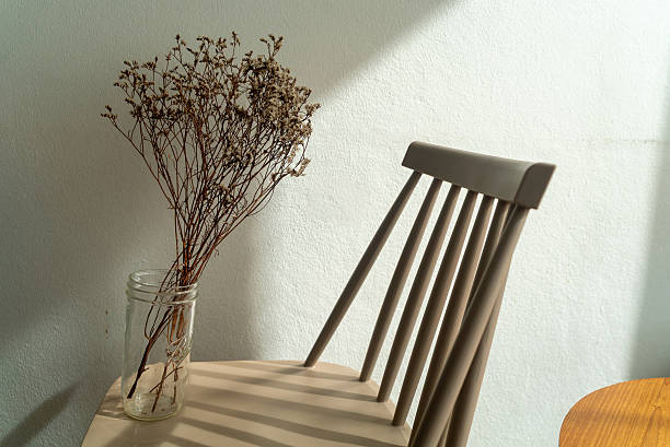 A minimalist scene featuring dried flowers in a jar placed on a simple chair, casting soft shadows under natural sunlight, perfect for a serene environment.
