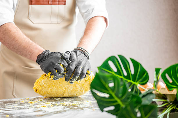 Chef kneading yellow dough on floured surface, wearing gloves and apron. Fresh ingredients and monstera plant in foreground. Culinary preparation in rustic kitchen setting.