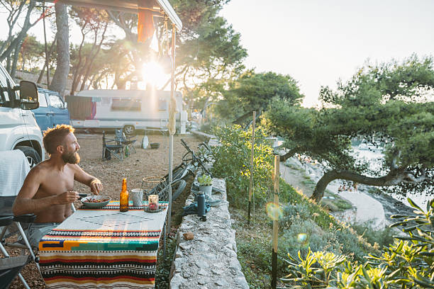 Bearded man eating vegan meal at outdoor campsite by the sea during sunset. Relaxed solo camper enjoying healthy food and nature. Camping lifestyle with scenic coastal view.