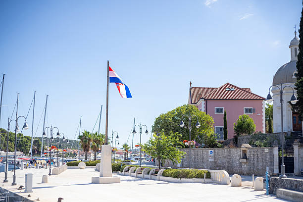 Croatian flag flying at harbor promenade with sailboats docked nearby. Pink historic house and church dome in background. Scenic coastal town with clear blue sky and Mediterranean vegetation.