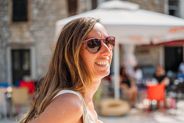 Happy woman with long hair wearing pink sunglasses and a white tank top outdoors. Smiling young adult enjoying sunny day in a casual urban setting. Portrait of a cheerful person outside.