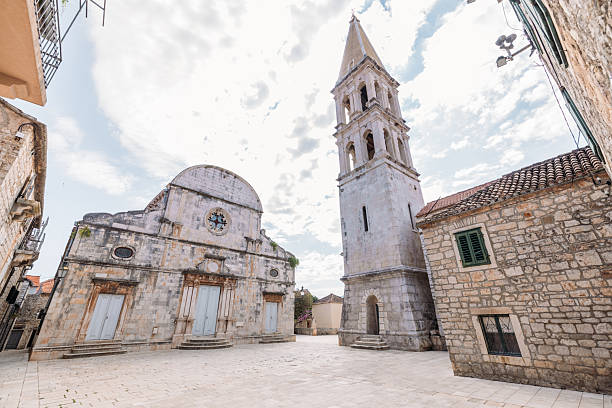 Old stone church with closed wooden doors and a tall bell tower under a partly cloudy sky. Historic architecture in a quiet town square with stone buildings and tiled roofs. European heritage site.