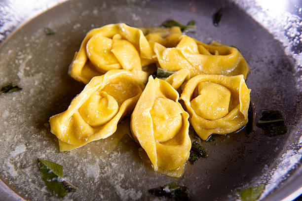A professional chef carefully shapes fresh, handmade tortellini on a floured wooden board, highlighting the traditional Italian culinary art. The image captures the meticulous process and skill required to create this iconic pasta, showcasing an authentic, rustic food preparation scene.