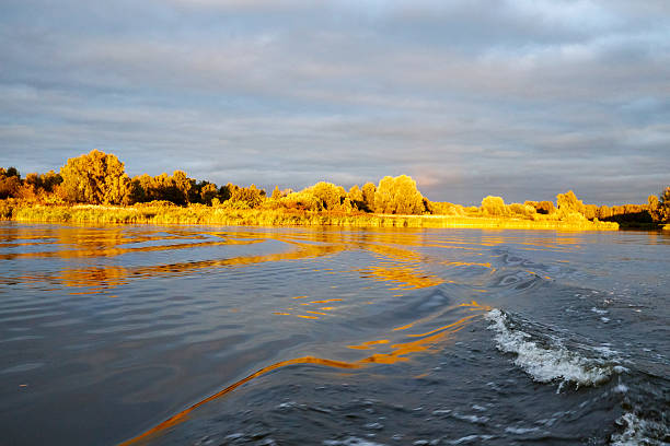 Golden sunset over river with dramatic clouds and bright reflections on water. Autumn trees glowing in warm evening light, creating a peaceful natural landscape with rippling waves