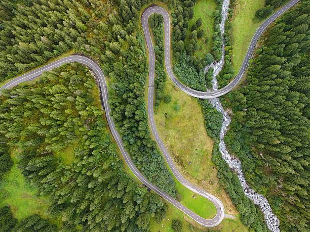 Drone view of a curvy mountain road snaking through dense green forest and alpine landscape, with a river running below.