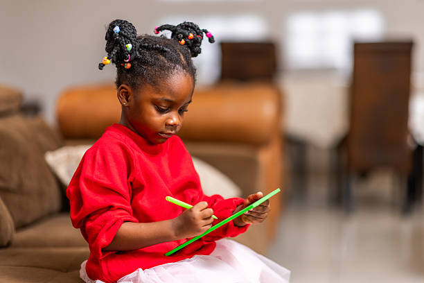 African girl child sitting on a couch using a tablet for homework, online learning, drawing, educational games, watching videos, and internet browsing.