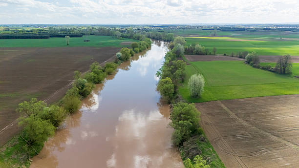 istock Aerial view of a winding river flowing through lush green fields with blooming trees under a partly cloudy sky. Ideal for nature and landscape themes. 2234366015