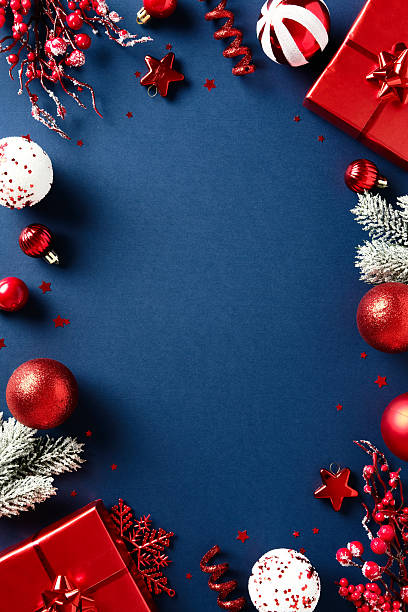 Vertical Christmas flat lay composition with red gift, frosted pine branches, and ornaments on dark blue background. Elegant seasonal design.