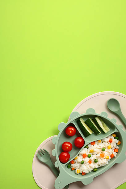 Playful dinosaur-shaped kids plate with rice, vegetables, and fresh greens, served with matching green cutlery on light green background, promoting healthy eating for children.