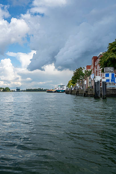 Dordrecht, Netherlands  5 September 2025: Old historic Dutch buildings rise along the riverfront Oude Maas beneath stormy skies, with inland shipping vessels and a cruise ship docked nearby.