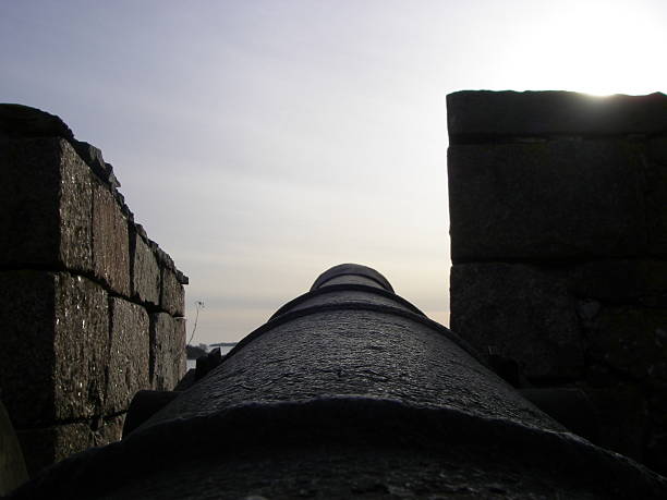Low-angle view down a dark, textured cannon barrel positioned between two stone fortress walls, silhouetted against a bright, hazy sky with a glimpse of sun.