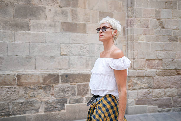 A woman wearing a white shirt and yellow plaid skirt stands in front of a brick wall. She is wearing sunglasses and has her hair pulled back