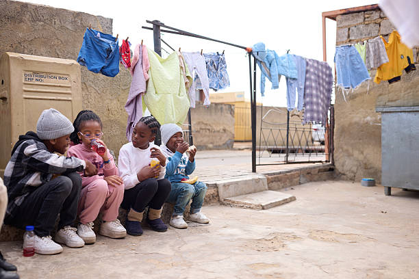 Children having snacks and refreshments while sitting next to each other. Siblings are communicating while spending leisure time in front yard. Clothes can be seen hanging on rope.