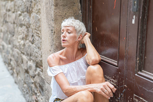 Young woman sits on a stone step in a quiet old town street while looking upward in natural daylight. The scene conveys calmness, confidence, and relaxed summer lifestyle.