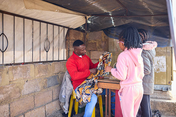 Tailor sitting on chair and measuring fabric at his shop. Man is talking with girls while holding cloth. Concept of small business.