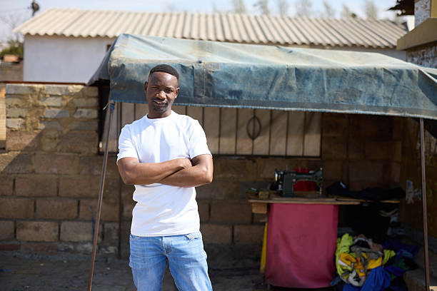 Portrait of male tailor posing with arms crossed in front of his shop. Man in white t-shirt at sunny day. He seems to be confident.
