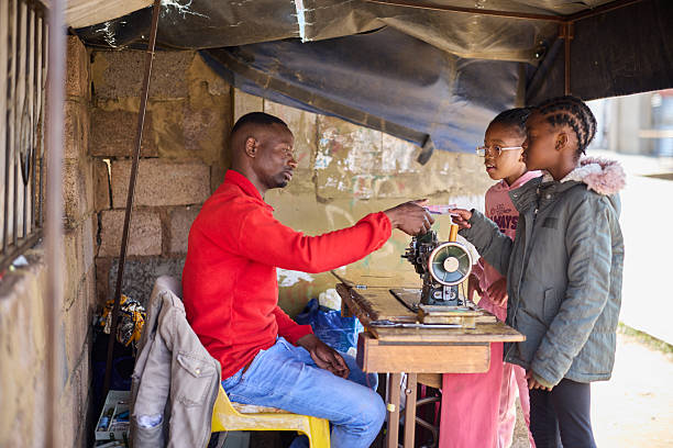 Girls are making payment at street side tailor shop. Tailor is taking money from customers. He is sitting on chair.
