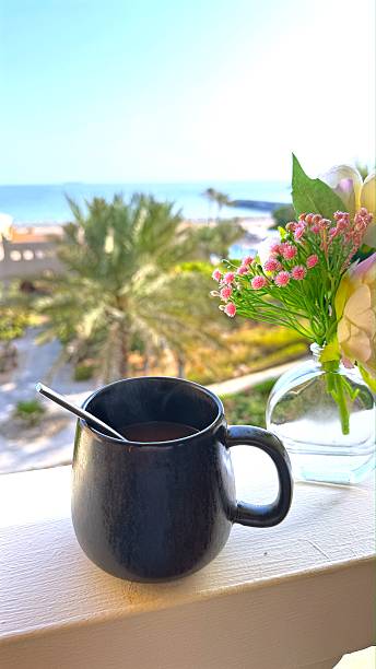 Steaming coffee cup resting on white balcony railing, tropical beach background with swaying palms and floral vase, embodying serene morning atmosphere