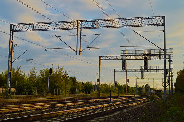 Overhead electric railway lines and signals silhouetted against a cloudy evening sky, surrounded by trees and infrastructure