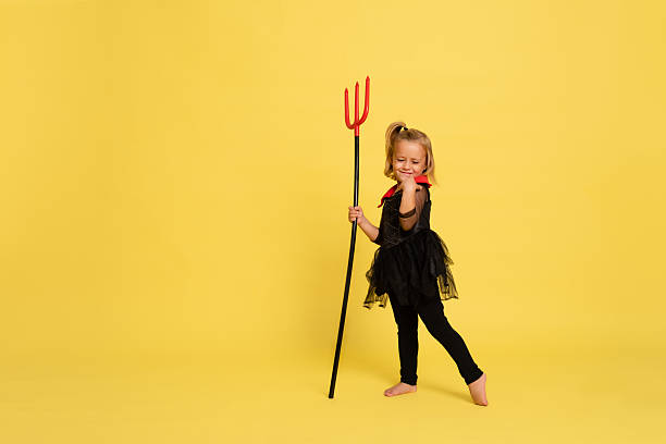 Playful child in black halloween costume with red collar smiling and holding trident on yellow studio background horizontal