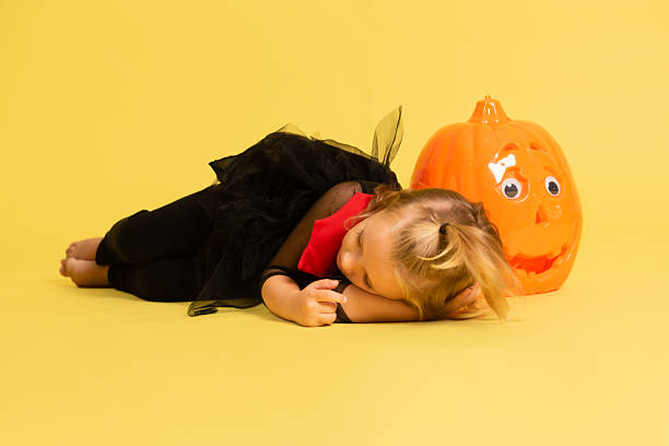 Girl in halloween outfit resting on yellow background with large orange pumpkin prop horizontal