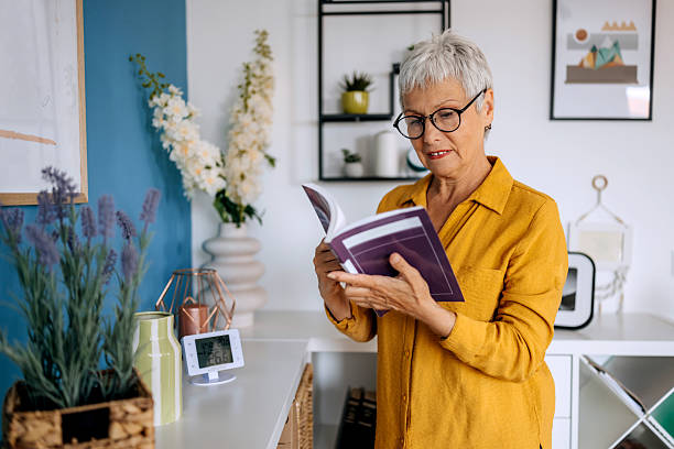 istock Senior woman reading book while standing near shelf at home 2234457991