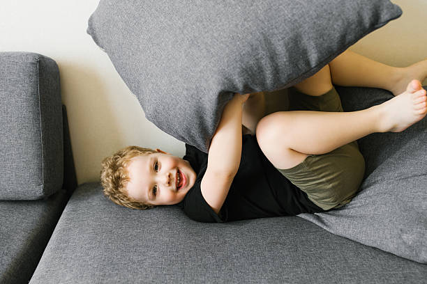A playful child lying on a gray sofa, laughing and hiding behind a large gray pillow.