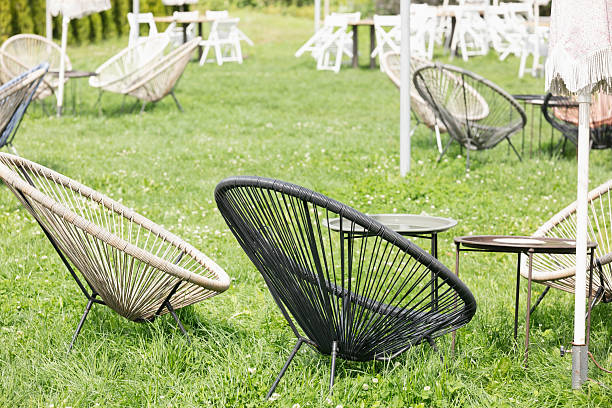 There are wicker chairs made of plastic in a row outdoors, against a background of green bushes. The cafe is closed, there are no visitors in the early morning. With space to copy. High quality photo