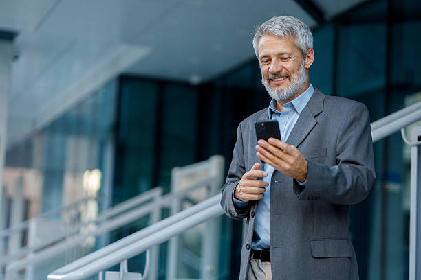 istock Confident mature businessman standing near modern office tower, smiling while using smartphone and embracing digital workplace communication 2234470895