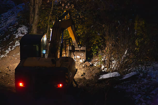 Excavator under headlights digs soil at night on construction site with trees and snow, nonstop construction work in winter period