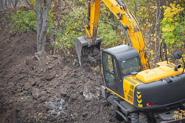 istock Excavator digs soil with bucket at construction site with trees under difficult working conditions. 2234475420