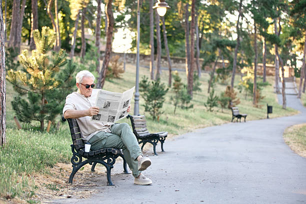 Elderly man wearing sunglasses, seated on a bench in a serene park, reading a newspaper amidst a backdrop of trees and greenery, enjoying a calm and relaxed outdoor afternoon.
