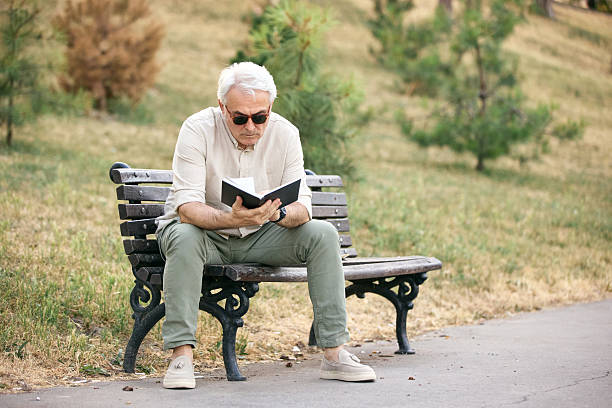 istock Elderly Man Reading a Book on a Bench in a Park 2234479614
